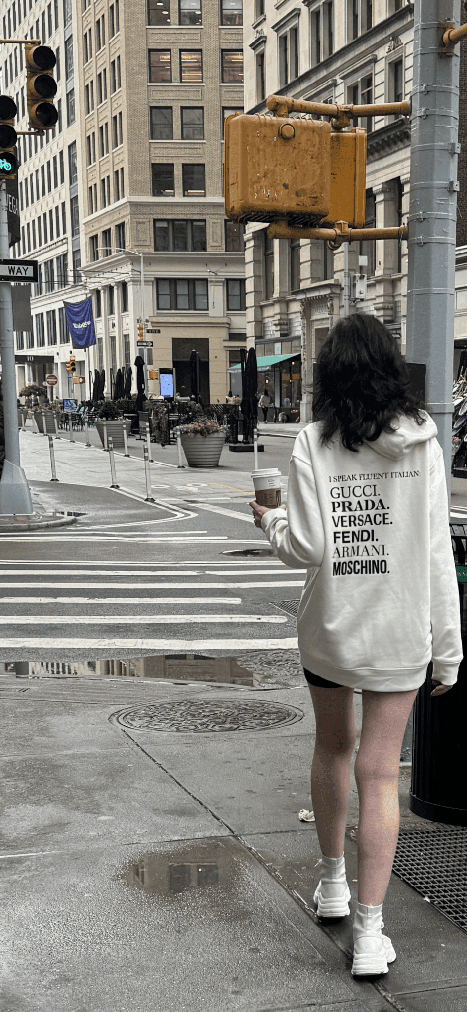 Sweat-shirt oversized unisexe ITALIAN LANGUAGE SWEAT blanc avec imprimé noir listant des marques italiennes de luxe, porté en ville. Style streetwear écoresponsable.